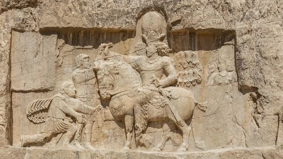 An ancient rock face relief at Naqsh-e Rostam shows Emperor Shapur on horseback compelling the captive Roman Emperor Valerian to kneel.